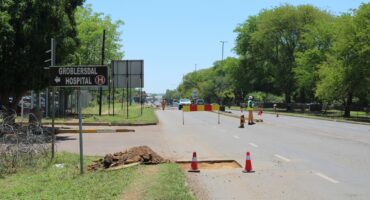 Road construction scene with a "Groblersdal Hospital" sign on the left. Traffic cones and barriers on road; workers in reflective vests managing traffic under clear skies.