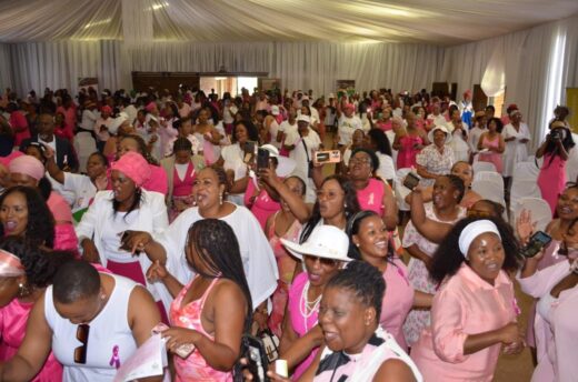 A large group of women wearing pink and white outfits gather in a decorated hall smiling and taking photos. The atmosphere is joyful and lively.