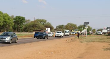 A stop-and-go point is visible on a busy road with cars and trucks under a clear blue sky, bordered by a dusty, grassy verge and lush green trees. A person walks by the roadside.