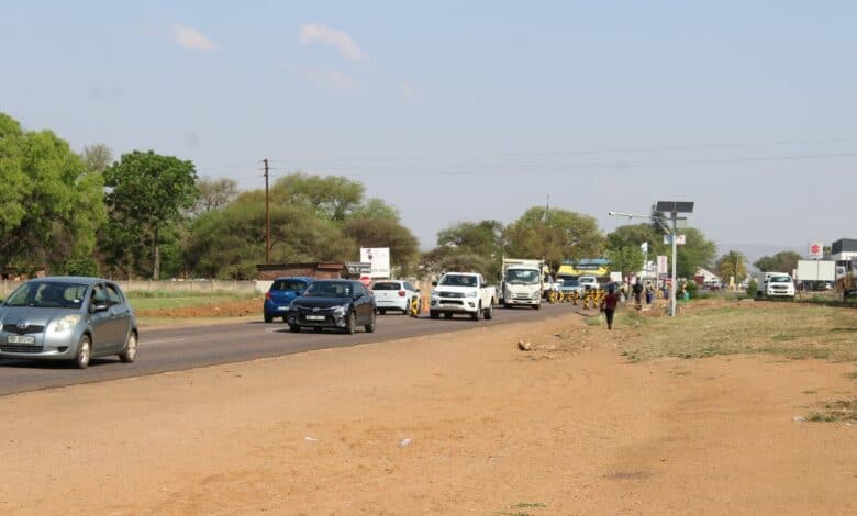A stop-and-go point is visible on a busy road with cars and trucks under a clear blue sky, bordered by a dusty, grassy verge and lush green trees. A person walks by the roadside.