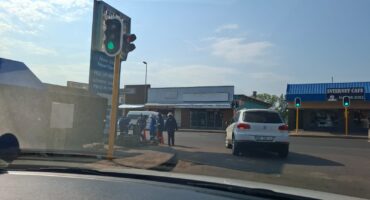 Street view at a traffic light intersection; green light, white SUV turning, municipal workers gathered near the intersection doing maintenance work, clear sky.