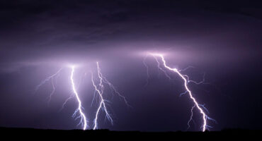 A vivid night sky is illuminated by bright, jagged lightning bolts striking the ground, conveying a sense of power and intensity against a dark horizon.