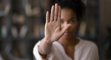 A person with an outstretched hand facing the camera, signaling stop. The background is blurred, and the mood conveys seriousness and assertiveness.