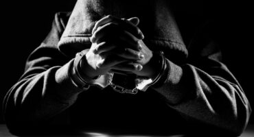 A hooded person sits in the dark with hands clasped, wearing handcuffs. Dramatic lighting creates a tense, somber mood, highlighting their predicament.