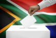A hand casts a vote into a ballot box, set against the backdrop of the South African flag. The image conveys a sense of civic duty and national pride.