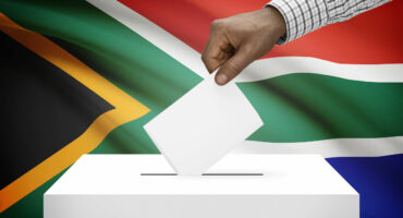A hand casts a vote into a ballot box, set against the backdrop of the South African flag. The image conveys a sense of civic duty and national pride.