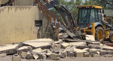 A yellow backhoe demolishes a beige wall, scattering rubble on the ground. Trees and a clear blue sky are visible in the background. The scene conveys destruction.