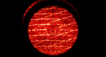A brightly lit red traffic light against a black background. The lens displays intricate patterns, conveying a sense of caution and urgency.