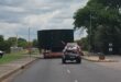 Truck transporting large green tank on a suburban road under cloudy skies. A small escort vehicle follows, emphasizing caution and safety.