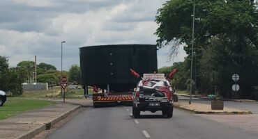 Truck transporting large green tank on a suburban road under cloudy skies. A small escort vehicle follows, emphasizing caution and safety.