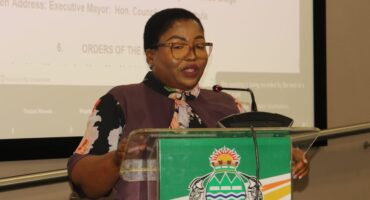 A woman with glasses speaks at a podium with a microphone, displaying the colorful emblem of the Sekhukhune District Municipality. The background shows a presentation slide, creating a formal tone.