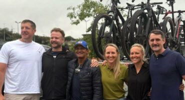 A group of six smiling people stands in front of a car with bikes on the rack. The overcast sky and casual attire suggest a relaxed, cheerful outing.