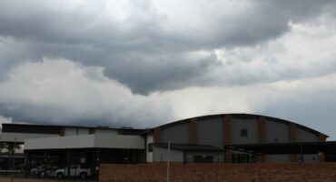 Overcast sky with dark, heavy clouds looming over a modern building with a curved roof and brick facade. The mood is ominous and foreboding.