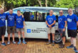 Six men in blue shirts stand smiling in front of a white van with "Eastvaal Motors" logo. Background shows trees and bicycles, creating a cheerful vibe.