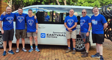 Six men in blue shirts stand smiling in front of a white van with "Eastvaal Motors" logo. Background shows trees and bicycles, creating a cheerful vibe.