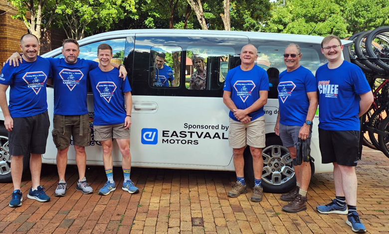 Six men in blue shirts stand smiling in front of a white van with "Eastvaal Motors" logo. Background shows trees and bicycles, creating a cheerful vibe.