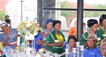A group of people in colorful traditional attire are gathered around a decorated table with flowers and glassware, exuding a festive and joyful atmosphere.