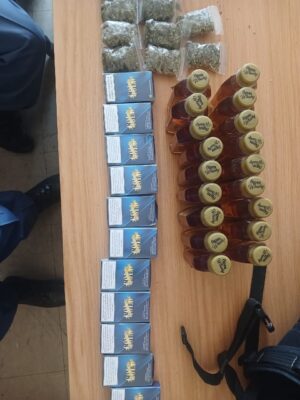 Packets of weed, blue cigarette packs, and small whiskey bottles arranged neatly on a wooden table. Two people partially visible beside the table.