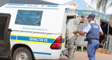 A police officer in uniform assists a person into a marked police vehicle labeled "Dennilton K9" in an urban setting with tents and signage nearby.