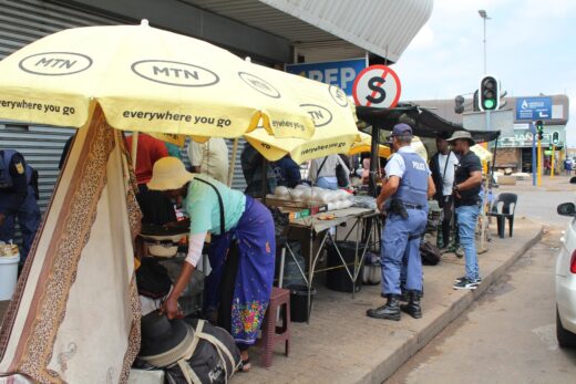 Street market scene with vendors under yellow umbrellas selling produce. Policeman in blue uniform nearby. Busy, bustling urban setting with a traffic light.