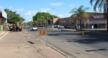 Sunny street scene showing light traffic, construction equipment, and workers. Palm trees and businesses line the road, creating a calm urban vibe.