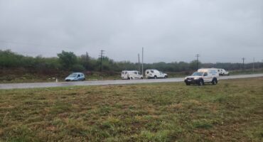 Three vehicles are parked on a wet roadside under a cloudy sky, including two white vans and a light blue car, creating a somber, rainy atmosphere.