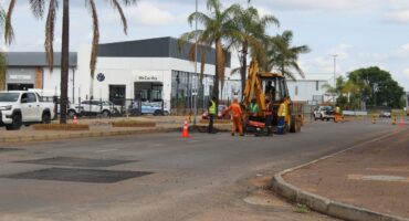 Roadwork scene on a city street with workers in orange uniforms near a backhoe. Traffic cones guide a passing white truck. Background features palm trees and a McCarthy car dealership.