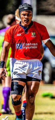 Rugby player in red jersey and white shorts walks on field. He wears a headgear and knee brace, embodying focus and determination. Bright, sunny day.
