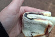 A hand holds a sandwich made of white bread with a visible piece of charred, blackened ham inside. The black poison can also be seen.