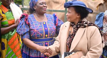 Two women in vibrant traditional attire shake hands outdoors, smiling warmly. One wears a colorful patterned dress, the other a beige coat and hat.