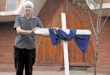Ds Wielie van Wyk stands beside a large white cross draped with a blue cloth, in front of wooden church doors. The mood is solemn and respectful.