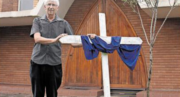 Ds Wielie van Wyk stands beside a large white cross draped with a blue cloth, in front of wooden church doors. The mood is solemn and respectful.