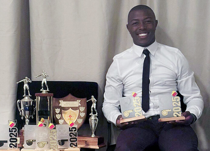 Excellent Mnkomo in a white shirt and tie smiles while seated, holding two "2025" awards. Beside him, several trophies and plaques are displayed.