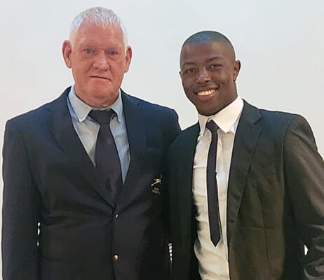 Steve Brits and Excellent Mnkomo in suits are smiling while posing together against a plain backdrop. The atmosphere is formal yet friendly, suggesting a special event.