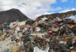 A large pile of mixed waste at a landfill, with debris like plastics and cardboard. A bird soars above, under a cloudy sky.