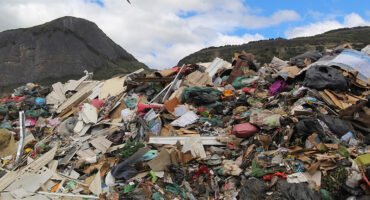 A large pile of mixed waste at a landfill, with debris like plastics and cardboard. A bird soars above, under a cloudy sky.