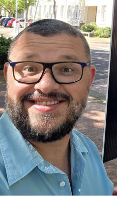 Smiling Franco Prinsloo with glasses and a beard wearing a light blue shirt, outdoors on a sunny day with trees, bushes, and buildings in the background.
