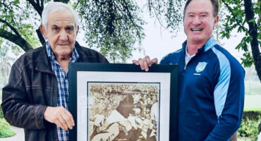 Two men stand outdoors, smiling, under a tree. They hold a framed vintage sports photograph. The scene conveys a sense of nostalgia and camaraderie.