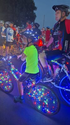 A group of cyclists at dusk, featuring a child and an adult on bikes adorned with colorful LED lights. The scene conveys excitement and community.