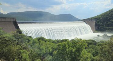 A large dam with water cascading over the edge, surrounded by lush green trees and rolling hills under a partly cloudy sky, conveying tranquility.
