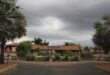 Entrance to "Royal Palm" estate with a roundabout and tropical plants. Overcast sky sets a somber tone. Solar panels are visible on a red-tiled roof.