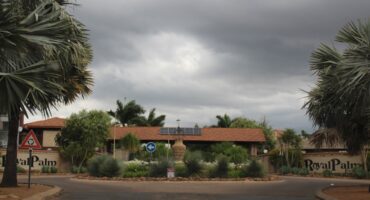 Entrance to "Royal Palm" estate with a roundabout and tropical plants. Overcast sky sets a somber tone. Solar panels are visible on a red-tiled roof.