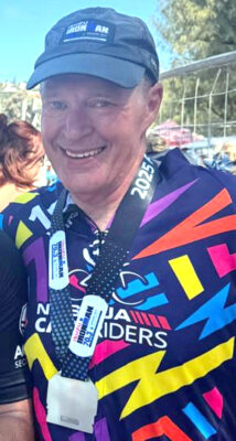 Smiling man in a colorful shirt and cap stands outdoors, wearing a finisher's medal. The image conveys a sense of achievement and joy.