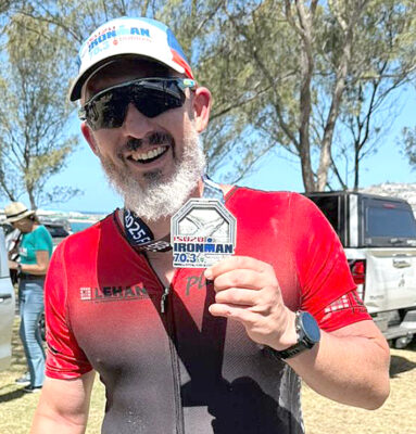 A man in a red athletic shirt smiles broadly, holding an Ironman 70.3 medal against a backdrop of trees and vehicles, exuding pride and joy.