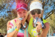 Two smiling women in athletic gear celebrate outdoors, biting medals. They wear visors and sunglasses, exuding joy and triumph amidst a sunny backdrop.