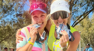 Two smiling women in athletic gear celebrate outdoors, biting medals. They wear visors and sunglasses, exuding joy and triumph amidst a sunny backdrop.