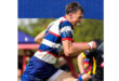 A rugby player in a blue, red, and white striped jersey runs energetically on a field. He appears focused and determined, with blurred teammates and greenery in the background.