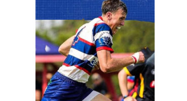 A rugby player in a blue, red, and white striped jersey runs energetically on a field. He appears focused and determined, with blurred teammates and greenery in the background.