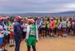 A diverse group of marathon runners in colorful attire and numbered bibs gathers at the starting line on a dirt path, with a scenic landscape in the background.