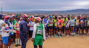 A diverse group of marathon runners in colorful attire and numbered bibs gathers at the starting line on a dirt path, with a scenic landscape in the background.